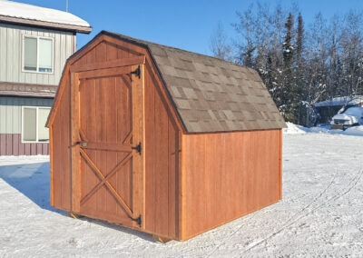 Exterior view of a Barn with a gambrel roof, cedar-colored siding, a single barn door, and brown shingle roofing, set in a snowy landscape near a building.