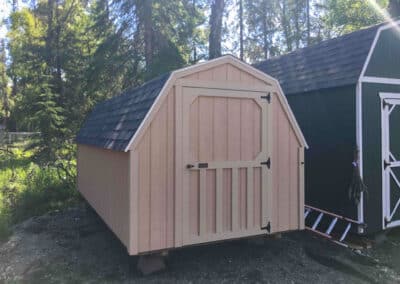 Primed Barn with white trim, black hardware, and a gambrel roof, positioned next to a dark green building in a wooded area.