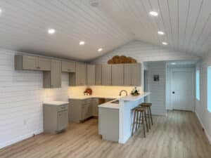 A modern kitchen in the Bighorn modular building featuring gray cabinetry, a white countertop, a breakfast bar with stools, shiplap walls, wood-style flooring, and recessed lighting.