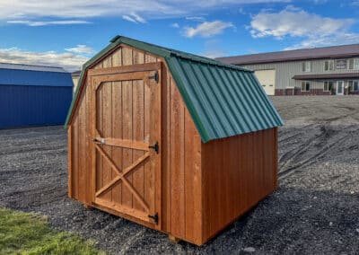 A small cedar-stained Barn with a gambrel roof, green metal roofing, and a single barn-style door, set on a gravel lot with other storage buildings in the background.