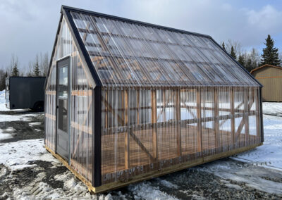 A wooden-framed greenhouse with clear polycarbonate panels, a black-framed door, and a gable roof, situated on a snowy gravel lot with surrounding outbuildings.