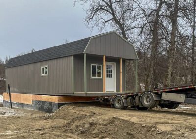 A lofted cabin with a gambrel roof being delivered on a flatbed trailer to a prepared foundation site surrounded by bare trees and winter conditions.