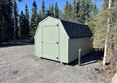 A green Barn with a gambrel roof and black shingles, featuring a single barn-style door, set on a gravel pad surrounded by a forest.