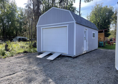 A gray Lofted Garage with a gambrel roof, white trim, white garage door with aluminum ramps, and a side entry door, set on a gravel pad surrounded by trees and nearby structures.