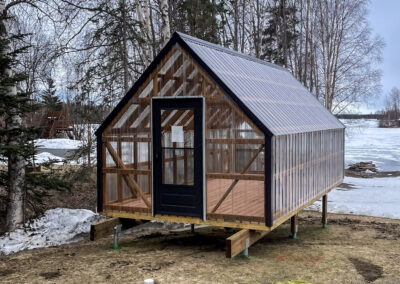 Greenhouse with a transparent design, wooden frame, and black-trimmed door, elevated on a lot near a snowy landscape.