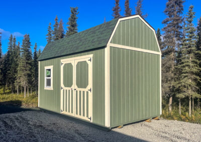 A green Lofted Barn with a gambrel roof, green shingles, white trim, and double barn-style doors, set on a gravel pad surrounded by a forest.