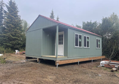 A green cabin with a gable roof, red metal roofing, white trim, and a covered porch, elevated on a Techno Metal Post foundation, set on a dirt lot surrounded by trees.