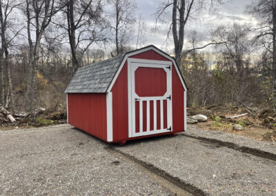 A red Barn with a gambrel roof, gray shingles, and white trim, featuring a single barn-style door, set on a gravel lot surrounded by bare trees.