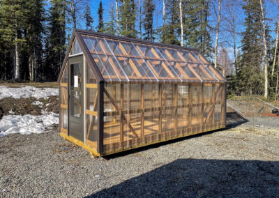 Greenhouse with a transparent polycarbonate exterior, wooden frame, and a single-door entry, set on a gravel lot surrounded by forest.