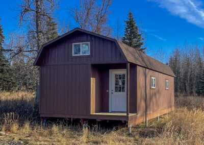 A brown Lofted Cabin with a gambrel roof, white door, and windows, elevated on a Techno Metal Post foundation, set in a grassy field with trees in the background.