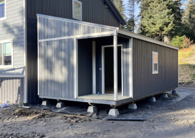 A gray Studio cabin with white trim, a covered porch, and energy-efficient windows, elevated on concrete pier blocks, set next to a modern house.