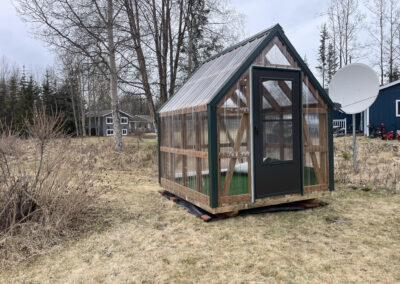 A small Greenhouse with transparent walls, green trim, and a dark brown door, set in a grassy yard with trees and houses in the background.