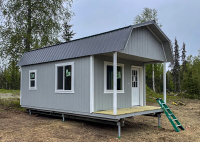 Light gray lofted cabin with a small porch, two windows, a white door, and a metal roof, set on a rural lot.