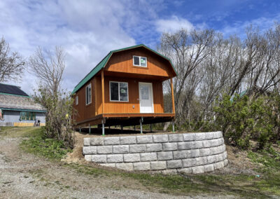 A brown Lofted Cabin with a gambrel roof and green metal trim, elevated on a Techno Metal Post foundation above a stone retaining wall, surrounded by trees and other buildings.