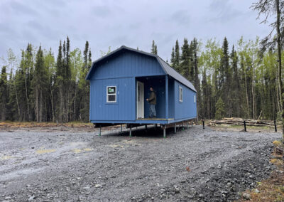 A blue Lofted Cabin with a gambrel roof and white trim, elevated on a Techno Metal Post foundation, set on a gravel clearing surrounded by a forest.