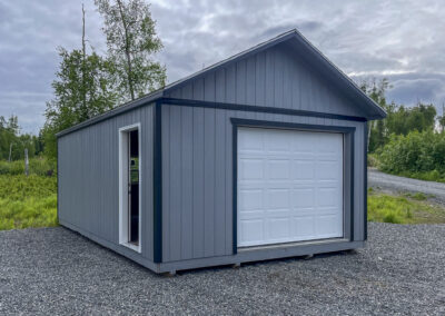 A gray Garage with a gable roof, white garage door, and side entry door, set on a gravel pad surrounded by greenery under a cloudy sky.