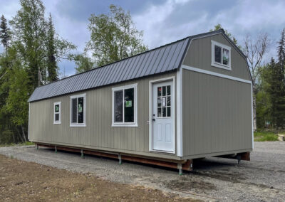 Taupe lofted barn with white trim, three large windows, a white door, and a metal roof, set in a wooded area.