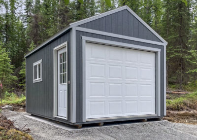 A gray Garage with a gable roof, white garage door, and side entry door, situated on a gravel pad surrounded by dense forest.