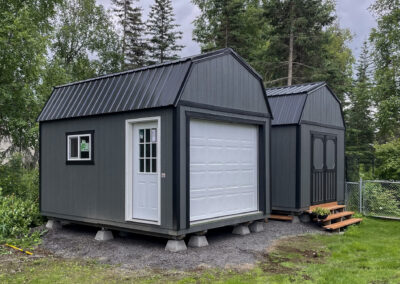 Two gray buildings, one Lofted Garage with a gambrel roof, metal roofing, a white garage door, and a side entry door, and one Barn with black trim and double doors, set on a gravel lot surrounded by greenery.