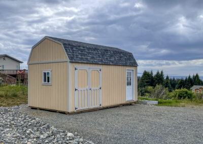 Lofted barn with white double doors, side window, and a shingle roof set in a residential area on a gravel lot.