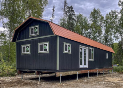 A black 16x52 Lofted Cabin with a gambrel roof, orange metal roofing, green trim, French doors, and multiple windows, installed on a Techno Metal Post foundation in a wooded area in Talkeetna, AK.