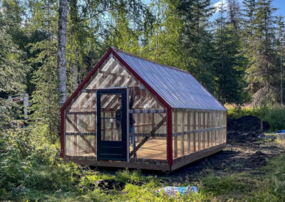 A wooden-framed Greenhouse with transparent polycarbonate panels, a red-trimmed gable roof, and a black door, set in a forested area.