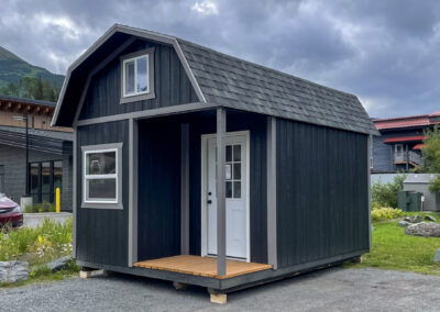 A black Lofted Cabin with a gambrel roof, gray trim, a small covered porch, and a white door, situated on a gravel area with a backdrop of buildings and mountains.