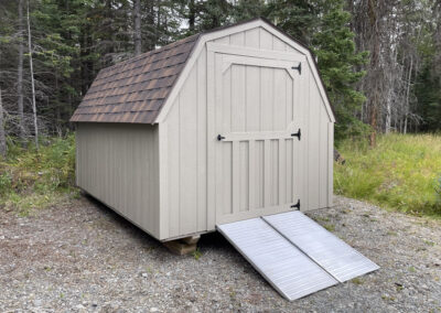 A beige Barn with a gambrel roof, brown shingle roofing, a single door, and a metal ramp, set on a gravel area surrounded by trees.