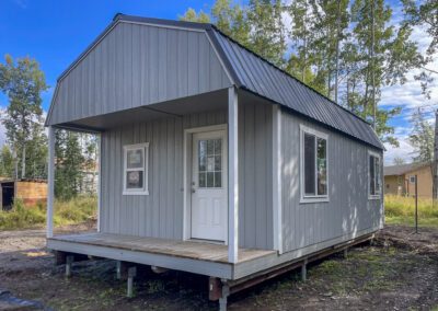 A gray Lofted Cabin with a gambrel roof, black metal roofing, a small covered porch, and white-trimmed windows, elevated on a raised foundation in a wooded area.