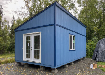 A blue Studio with a single-slope roof, French doors, white trim, and a side window, elevated on concrete piers, surrounded by trees.