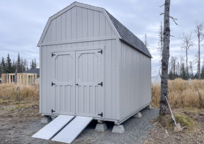 A gray Lofted Barn with a gambrel roof, shingle roofing, double barn doors, and two metal ramps, elevated on concrete piers in a rural setting.