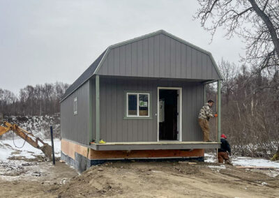 A Lofted Cabin with a gambrel roof being adjusted on a foundation by workers during winter, with a backhoe visible in the background.