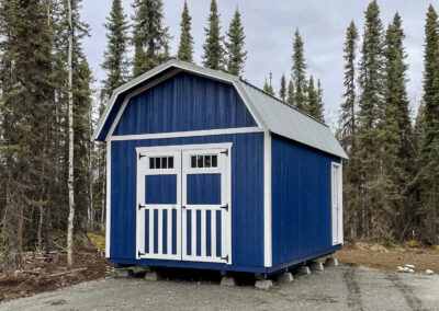 A blue Lofted Barn with a white metal gambrel roof, white trim, and double doors with decorative accents, elevated on concrete piers in a wooded area.