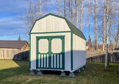 White lofted barn with green trim, double barn doors, and a green shingle roof, elevated on concrete blocks in a backyard surrounded by trees.