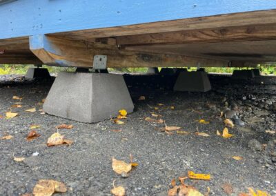 Close-up of a building foundation supported by concrete pier blocks and wooden skids, resting on a gravel surface scattered with autumn leaves.