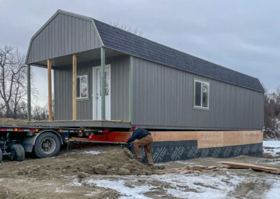 A Lofted Cabin with a gambrel roof being positioned on a crawlspace foundation by a worker, with a flatbed trailer still attached, during snowy winter conditions.