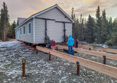 A beige building being maneuvered into place on a prepared foundation with wooden beams by a worker operating a Mule 747T, set in a snowy forest clearing at sunset.